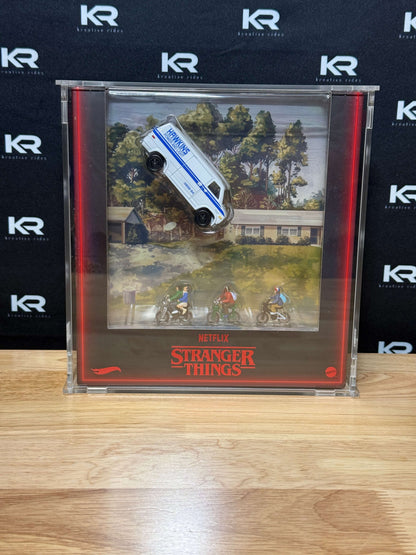 A Stranger Things collectible display inside a clear acrylic case, featuring a white and blue marked police car mounted above an illustrated scene with three riders on motorcycles and the red Stranger Things title card. The case sits on a light wood surface against a black branded backdrop.