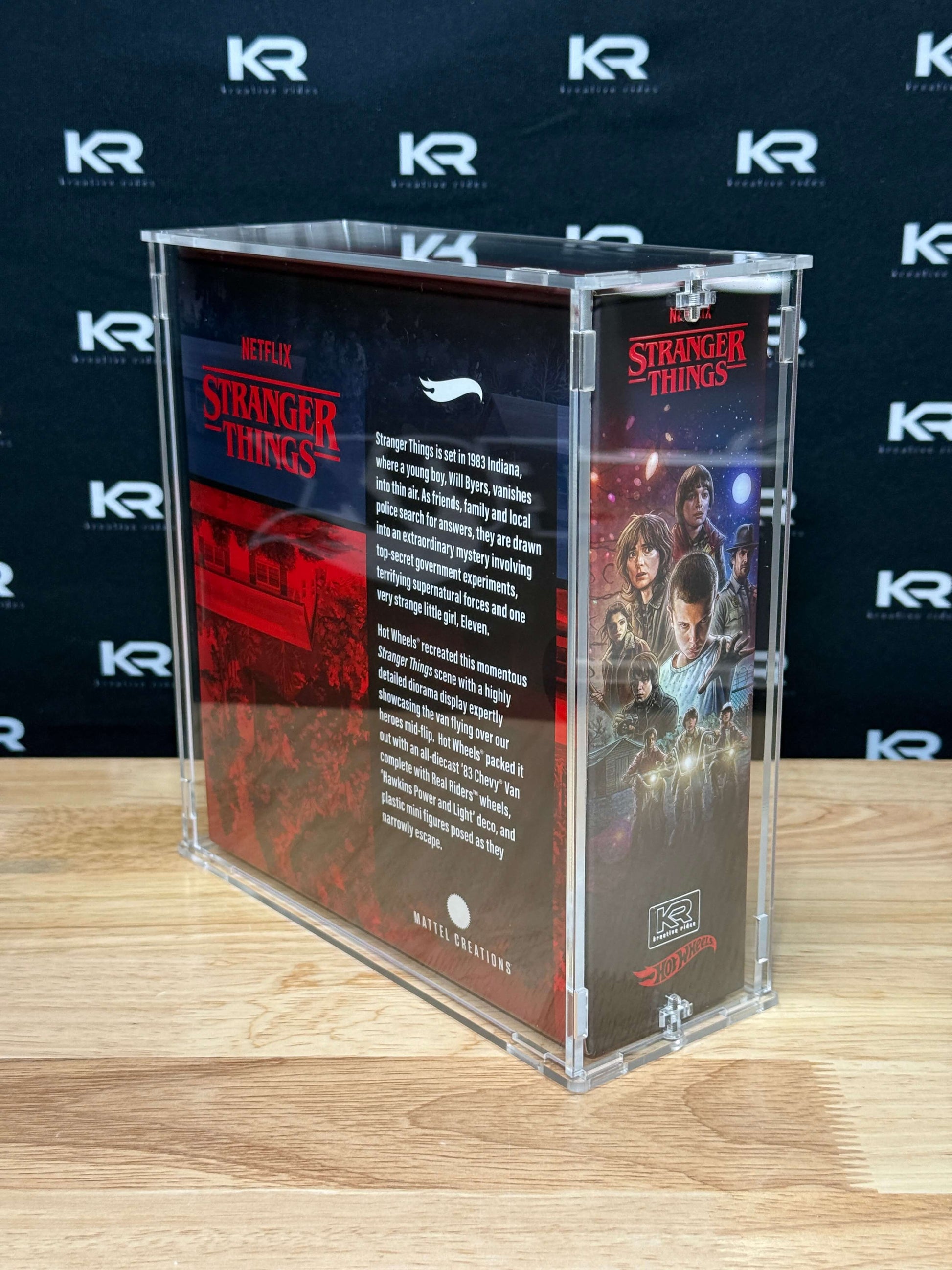 A clear acrylic display case containing Stranger Things merchandise, standing on a light wood tabletop against a dark branded backdrop. The case is transparent with printed Stranger Things artwork and text panels visible inside.