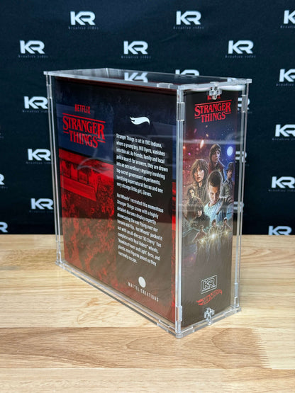 A clear acrylic display case containing Stranger Things merchandise, standing on a light wood tabletop against a dark branded backdrop. The case is transparent with printed Stranger Things artwork and text panels visible inside.