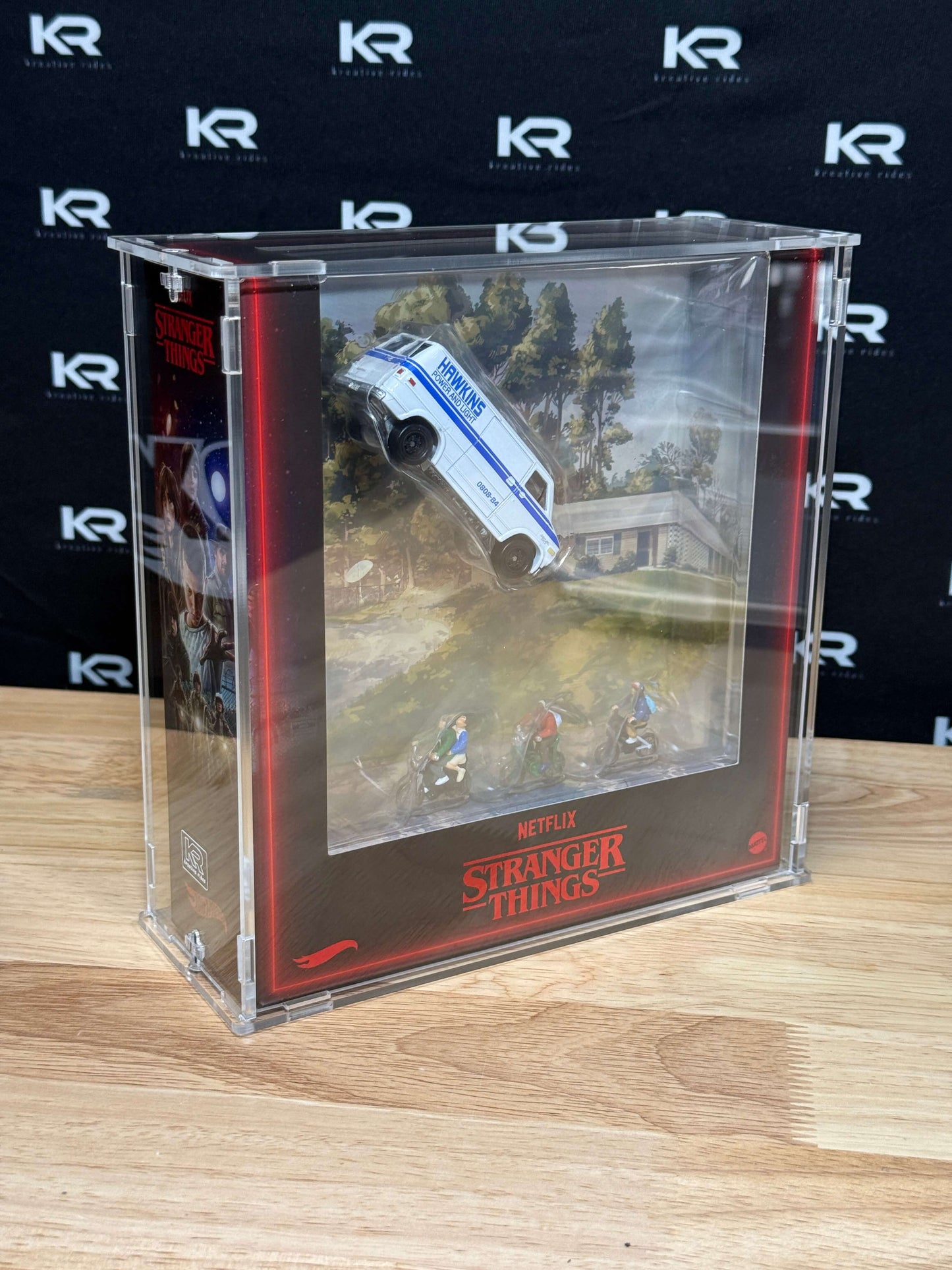 A clear acrylic display case containing a Stranger Things themed miniature scene with a white and blue vehicle and small figures, set on a wooden surface against a black branded backdrop.