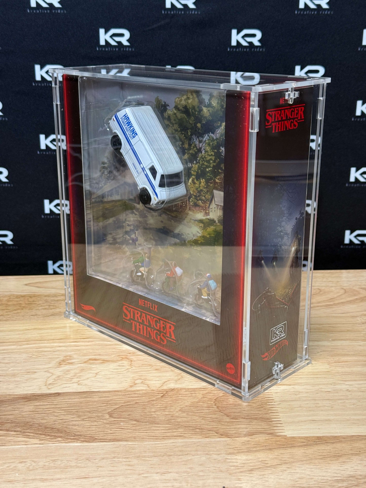 A clear acrylic display case containing a miniature Stranger Things scene with a white van and small figures, set on a wooden surface against a black branded backdrop.
