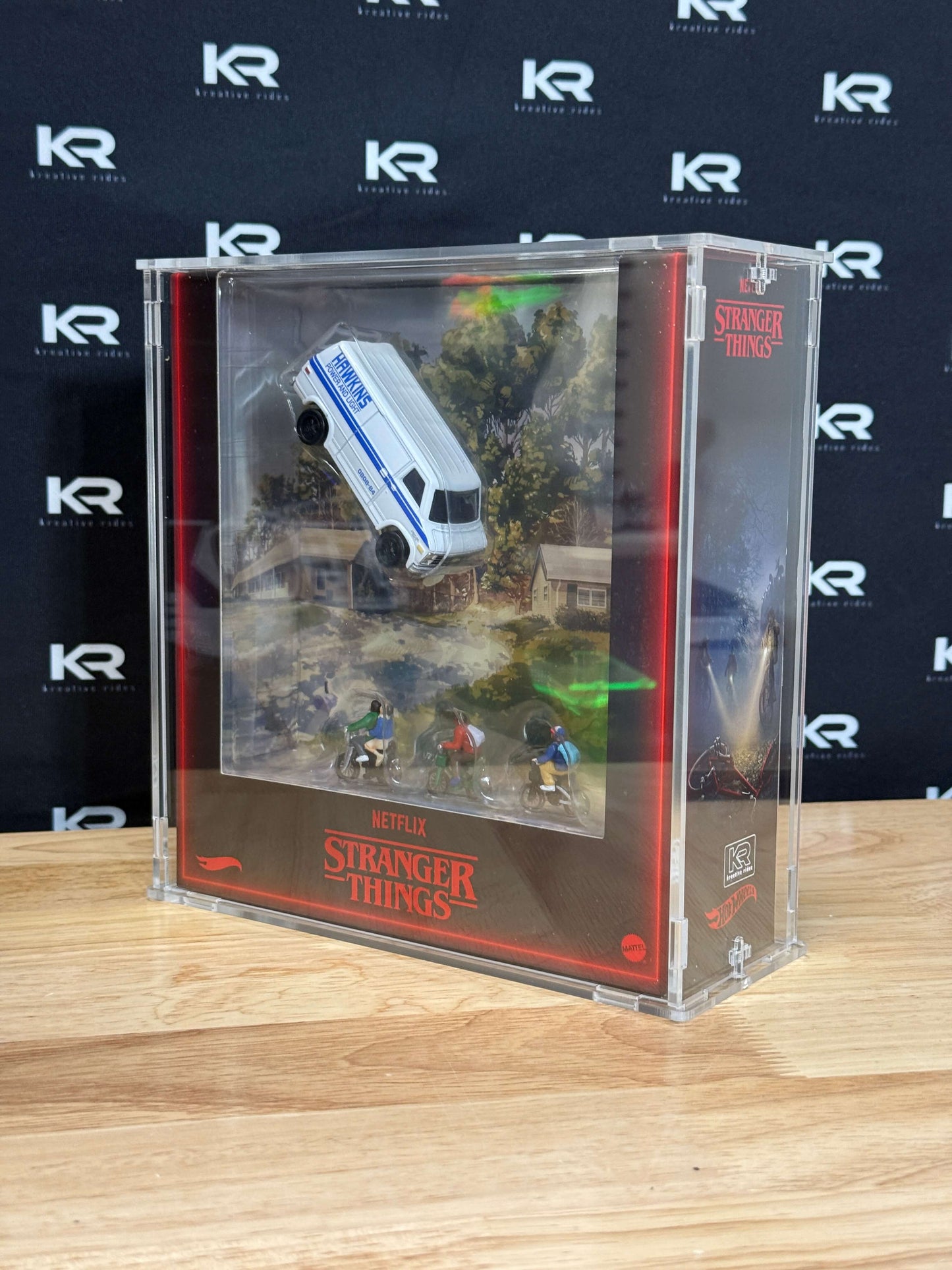 A clear acrylic display case containing a miniature Stranger Things scene with a white and blue van, small character figures, and printed scenery, set on a wooden surface against a branded black backdrop.