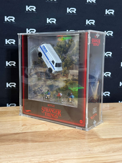 A clear acrylic display case containing a miniature Stranger Things scene with a white and blue van, small character figures, and printed scenery, set on a wooden surface against a branded black backdrop.
